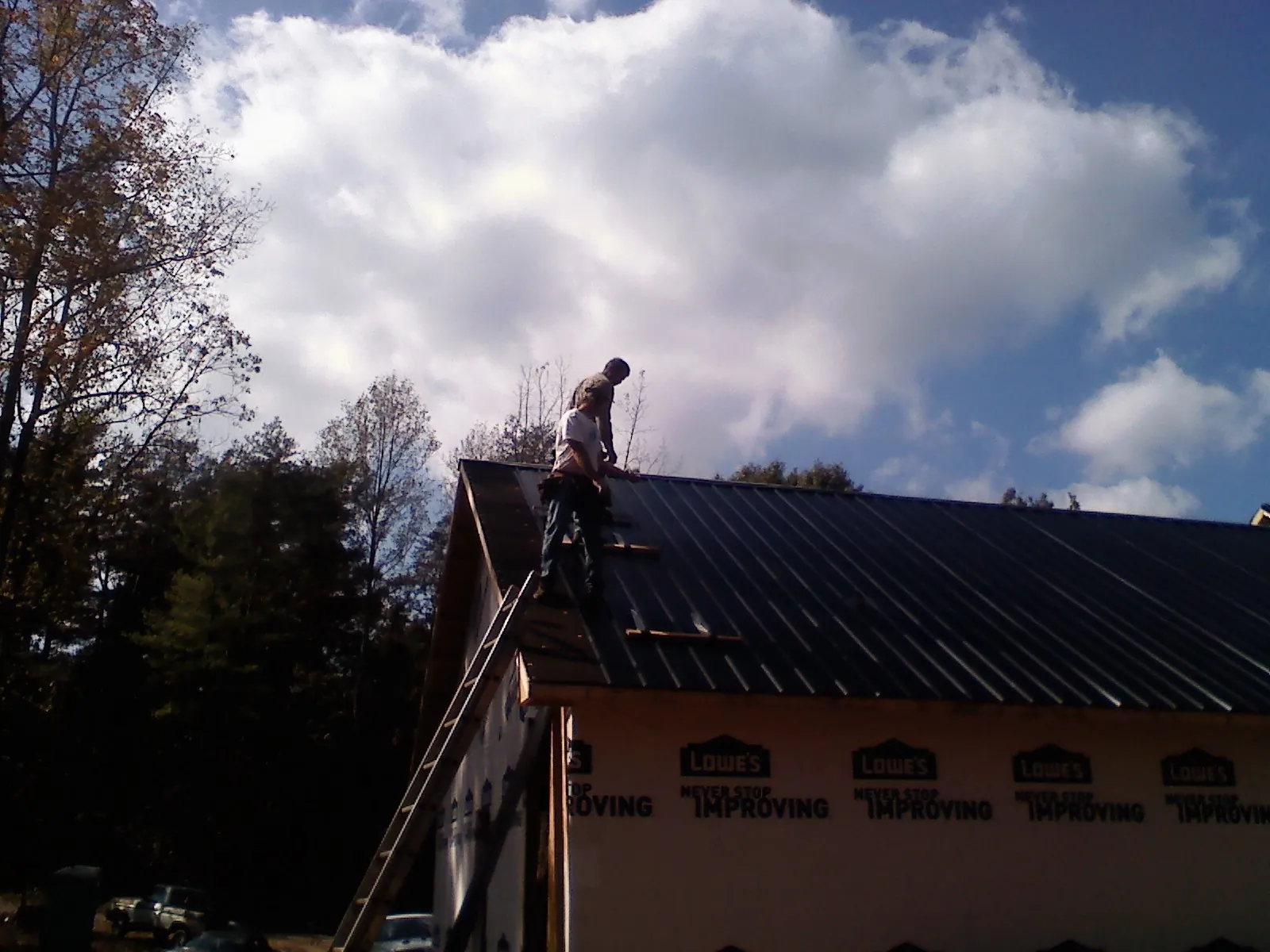 Roofer fastening dark metal panels high against a cloudy sky, from a Roof Inspection job in Wilsall